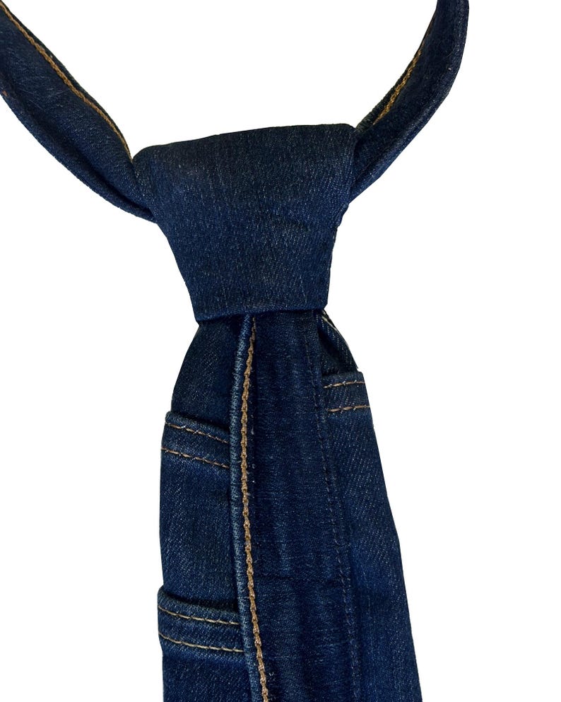 K&ouml;nnte beinhalten: Eine dunkelblaue Jeans-Krawatte mit sichtbaren N&auml;hten und Taschen-Details. Die Krawatte ist oben geknotet und zeigt die Textur und Konstruktion des Jeansstoffs. Das Design ahmt das Aussehen einer Jeans nach.