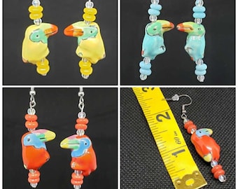 May include: A set of three pairs of toucan earrings. Each pair features a different color toucan: yellow, blue, and red. The toucans are made of ceramic and have painted details. The earrings are strung with clear and colored beads.
