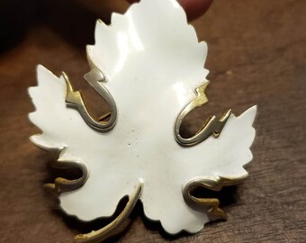 May include: A white enamel leaf brooch with gold accents. The leaf has a detailed design and is a vintage style.