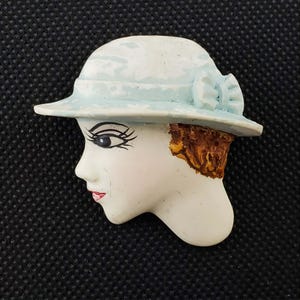 May include: A vintage-style brooch featuring a woman's profile. The brooch depicts a woman wearing a light blue hat with a bow. Her face is painted with detailed features, including dark eyelashes and red lips. The hair is brown.