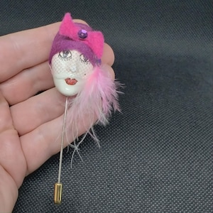 May include: A vintage-style hat pin featuring a painted face with red lips and a purple and pink hat. The hat is adorned with a pink bow and a purple gem. Pink feathers cascade from the side. The pin is silver-toned with a gold-toned base.