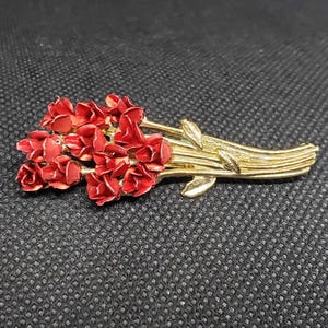 May include: A gold-toned brooch featuring a bouquet of red roses. The roses are clustered together, with detailed petals. The stems are gold, and the brooch has a classic, elegant design. The background is a textured black surface.