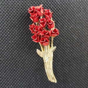 May include: A gold-toned brooch featuring a bouquet of red roses. The roses are clustered together and have a realistic appearance. The brooch is a delicate and elegant piece of jewelry.