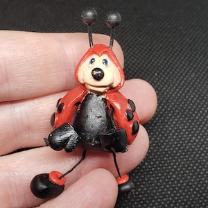 May include: A whimsical ladybug figurine with a red shell, black spots, and a black body. The ladybug has a smiling face with blue eyes and black antennae. Its legs are black with red accents.