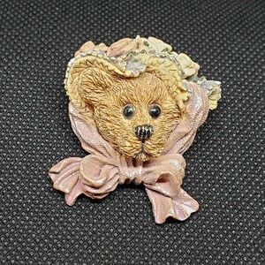 May include: A detailed brooch featuring a teddy bear wearing a hat adorned with flowers and a pink scarf tied in a bow. The bear has a textured fur appearance with black eyes and nose. The brooch is made of a light brown material.