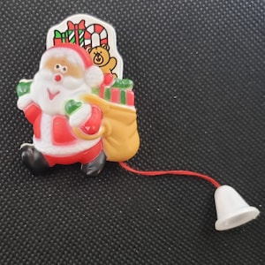 May include: A plastic Santa Claus pin featuring a red and white suit, carrying a yellow sack filled with green and red gifts. A small brown teddy bear is visible behind him. A thin red string hangs from the bottom, attached to a white bell.