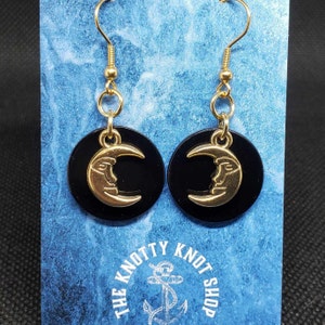 May include: Gold crescent moon earrings with a face on the moon, hanging from black round discs. The earrings are on a blue background with the text "The Knotty Knot Shop" and an anchor.