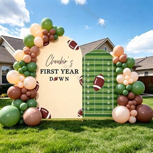 Pode incluir: Um fundo de festa com tema de futebol americano composto por dois painéis arqueados decorados com uma guirlanda de balões em tons de verde-oliva, castanho e creme. O painel esquerdo exibe o texto 'Choubin's FIRST YEAR DOWN' e o direito um campo.