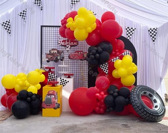 May include: A Cars-themed birthday party decoration with red, yellow, and black balloons, a black grid backdrop with the name "Fabian" and the number "2", a red and yellow toy truck, and a black tire.