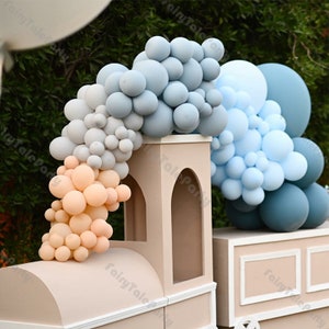 Puede incluir: Un telón de fondo de tren de color marrón claro decorado con un arco de globos. El arco está hecho de globos de color azul claro, gris y melocotón. Los globos están dispuestos en forma de cascada.