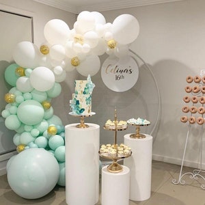 May include: A white and mint green balloon arch with gold confetti, a white cake with blue and green floral designs, and white cake stands with gold accents. A white round backdrop with a gold mesh design and the text "Celina's 16th" in gold lettering.