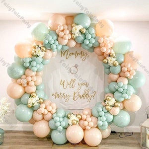May include: A balloon arch made of peach and light green balloons with gold accents. The arch is decorated with white flowers and a sign that says "Mommy, Will you marry Daddy?"