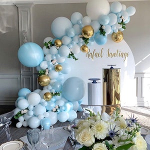 May include: A white round backdrop with the name "Rafael Santiago" written in gold lettering. The backdrop is decorated with a garland of blue and white balloons, gold balloons, and greenery.