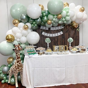 May include: A green, white, and gold balloon arch with the words "God Bless Leonardo" and "Leo" in gold letters. A stuffed giraffe stands in front of the arch. The arch is in front of a wooden wall with a white table and a dessert display.