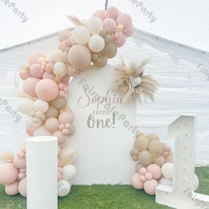 May include: A white backdrop with the text "Sophia turns one!" decorated with a balloon arch of white, pink, and beige balloons. The arch is accented with dried pampas grass. A white, illuminated number one stands to the right of the backdrop.