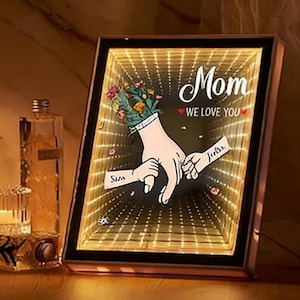 Peut inclure: Cadre lumineux avec l'inscription "Mom WE LOVE YOU" et un dessin de mains se tenant la main. L'œuvre présente un effet miroir infini et un bouquet floral. Les noms "Sim" et "Jada" sont également visibles.