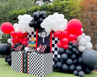 May include: A black, white, and red balloon arch with the text "Curtis 5" on a black and white checkered backdrop. A table with a checkered tablecloth is decorated with black and white checkered boxes and a red and yellow race car cake.