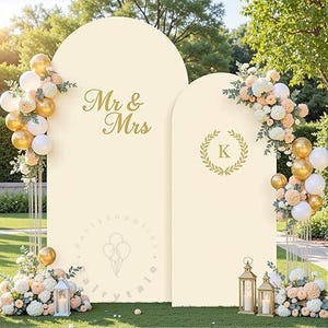 May include: Two cream-colored arched backdrop panels featuring gold text. The taller panel displays "Mr & Mrs" and the shorter panel features a letter "K" inside a laurel wreath. Both are decorated with clusters of gold and white balloons and flowers.