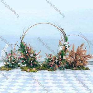 May include: Three gold metal hoops decorated with dried pampas grass, greenery, and flowers. The hoops are arranged in a staggered formation on a white and gray tiled floor.