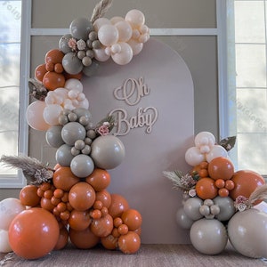 Op de afbeelding: Een beige boog achtergrond met de tekst "Oh Baby" in een lichtbruine lettertype. De achtergrond is versierd met een guirlande van ballonnen in tinten wit, grijs en oranje.