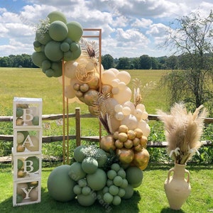 May include: A green, gold, and cream balloon arch with a sign that says "Baby" in gold letters. There are also pampas grass and a clear box with the letters "BABY" in white.