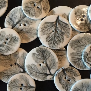 May include: A collection of round, white ceramic buttons with a leaf design in shades of grey. The buttons have two holes each.