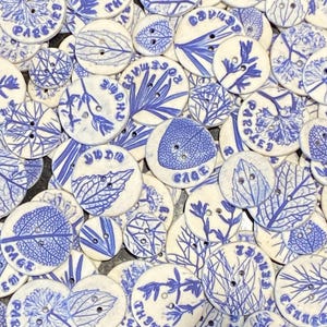May include: A selection of off-white ceramic buttons with blue botanical patterns and text. The buttons display diverse leaf and plant illustrations, with words such as "Sage," "Rosemary," and "Parsley" printed on them. Each button has two holes for sewing.