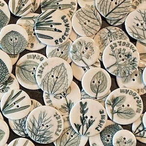 May include: A collection of round ceramic buttons featuring various botanical illustrations, including rosemary, thyme, mint, parsley, and sage. The buttons are white with green and black details.