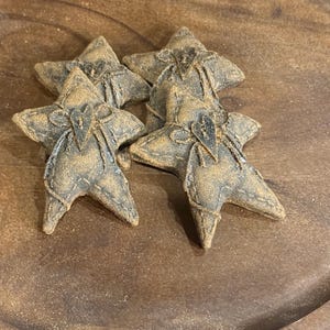 May include: Four star-shaped fabric ornaments in a muted dark brown and tan color palette. Each star features visible stitched edges, a central heart-shaped cutout, and a decorative twine bow tied around the center point of the star shape.