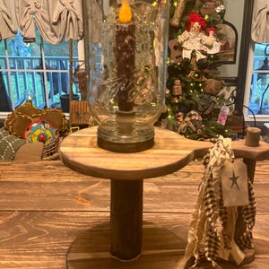 May include: A clear glass mason jar with a dark brown textured candle featuring a yellow flame light, resting on a round wooden pedestal stand. The stand includes a decorative fabric tassel with a small tag displaying a black star graphic.