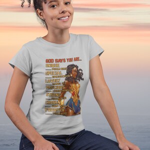 Puede incluir: Una camiseta blanca con un gr&aacute;fico de una mujer de piel morena y cabello rizado que lleva un disfraz de Wonder Woman. El texto de la camiseta dice "GOD SAYS YOU ARE... UNIQUE PSALM 139:13 SPECIAL EPHESIANS 2:10 LOVELY DANIEL 12:3 PRECIOUS 1 CORINTHIANS 6:20 STRONG PSALM 18:35 CHOSEN JOHN 15:16 FORGIVEN PSALM 103:12"
