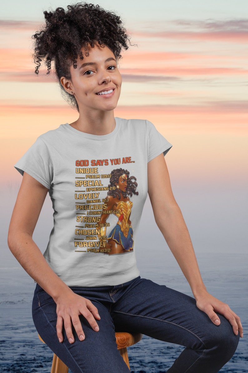 Puede incluir: Una camiseta blanca con un gr&aacute;fico de una mujer de piel morena y cabello rizado que lleva un disfraz de Wonder Woman. El texto de la camiseta dice "GOD SAYS YOU ARE... UNIQUE PSALM 139:13 SPECIAL EPHESIANS 2:10 LOVELY DANIEL 12:3 PRECIOUS 1 CORINTHIANS 6:20 STRONG PSALM 18:35 CHOSEN JOHN 15:16 FORGIVEN PSALM 103:12"
