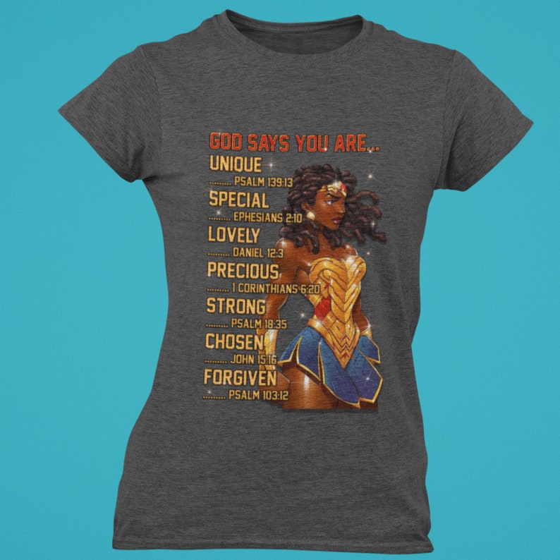 Puede incluir: Una camiseta gris con un gr&aacute;fico de una mujer de piel morena y cabello rizado que lleva un disfraz de Wonder Woman. El texto "GOD SAYS YOU ARE..." est&aacute; impreso en la camiseta con una lista de palabras y vers&iacute;culos b&iacute;blicos debajo.