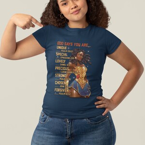 Puede incluir: Una camiseta azul marino con un gr&aacute;fico de una mujer con un traje dorado de Wonder Woman. El texto "GOD SAYS YOU ARE..." est&aacute; impreso en la camiseta con una lista de atributos positivos y vers&iacute;culos b&iacute;blicos correspondientes.