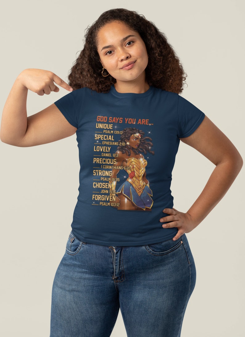 Puede incluir: Una camiseta azul marino con un gr&aacute;fico de una mujer con un traje dorado de Wonder Woman. El texto "GOD SAYS YOU ARE..." est&aacute; impreso en la camiseta con una lista de atributos positivos y vers&iacute;culos b&iacute;blicos correspondientes.