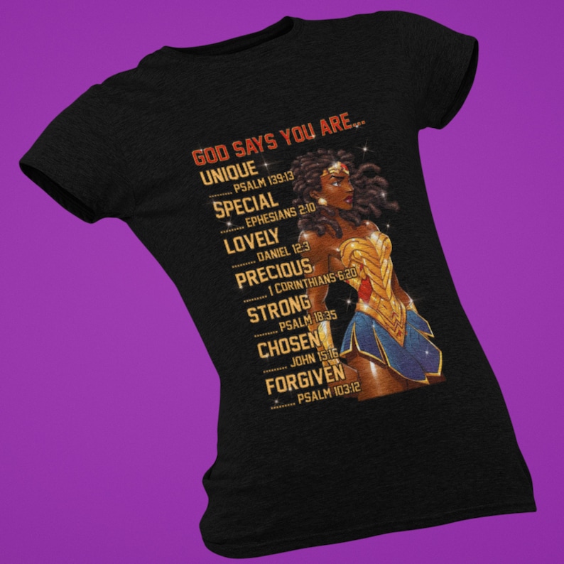 Puede incluir: Camiseta negra con un gr&aacute;fico de una mujer de piel morena y cabello rizado que lleva un disfraz de Wonder Woman. El texto "GOD SAYS YOU ARE..." est&aacute; impreso en la camiseta con una lista de palabras y vers&iacute;culos b&iacute;blicos.