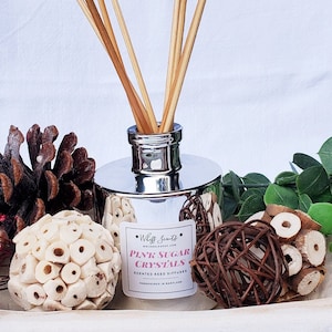 May include: A silver reed diffuser with wooden reeds, labeled "Pink Sugar Crystals." The image includes decorative elements like a pinecone, a white decorative ball, and a woven sphere, creating a cozy, aromatic ambiance.
