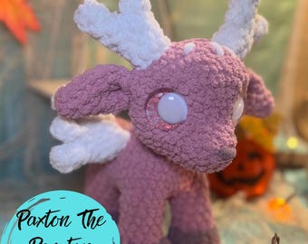 May include: A handmade crochet Peryton plushie in shades of pink and gray, with white antlers and wings. The plushie has large, round eyes and is labeled "Paxton The Peryton By Crochet With Cate". The text "PDF ONLY" is in the top left corner.