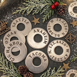 May include: A collection of circular metal washers stamped with various city names, coordinates, and dates. The washers are arranged among small decorative metal sleighs, gold stars, pine sprigs, red berries, and a string of warm white fairy lights.