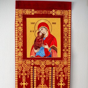Könnte beinhalten: Ein rotes Samtbanner mit goldener Stickerei, das eine religiöse Ikone der Heiligen Anna mit der Jungfrau Maria und dem Jesuskind darstellt. Das Banner hat drei Quasten und den Text "Gott ist mit uns" oben.