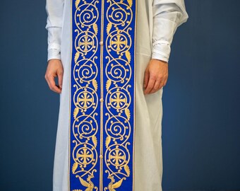 Pode incluir: Uma vestimenta litúrgica branca com uma estola bordada em azul e dourado. A estola apresenta desenhos repetidos de cruzes e pergaminhos dourados sobre um fundo azul. A bainha inferior da vestimenta tem uma borda bordada em dourado.