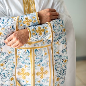 Może przedstawiać: Biała szata religijna ze złotym haftem wokół kołnierza i mankietów. Mankiety i przedni panel mają szczegółowy wzór niebieskich i złotych wzorów kwiatowych i krzyży. Szata jest noszona na beżowej bieliźnie.