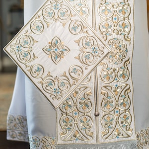 May include: White liturgical vestment with intricate gold and blue embroidery. The vestment features a square panel with a floral design and a fringe trim.