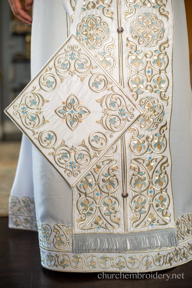 May include: White liturgical vestment with intricate gold and blue embroidery. The vestment features a square panel with a floral design and a fringe trim.