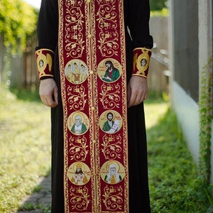 Pode incluir: Uma veste lit&uacute;rgica vermelha com bordados dourados e v&aacute;rios &iacute;cones circulares de santos. A veste tem uma franja.