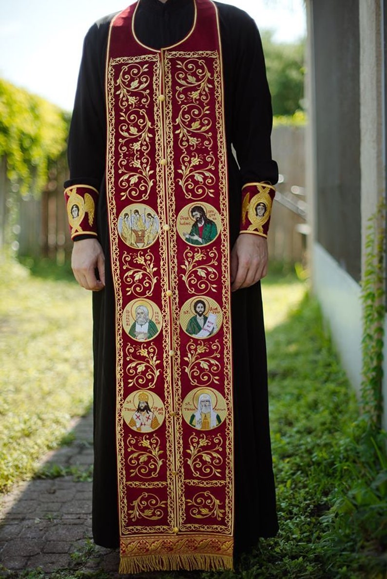 Pode incluir: Uma veste lit&uacute;rgica vermelha com bordados dourados e v&aacute;rios &iacute;cones circulares de santos. A veste tem uma franja.