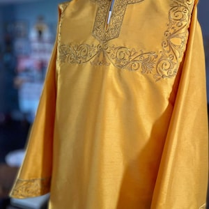 May include: A long-sleeved, gold-colored tunic with intricate gold embroidery around the neckline and cuffs. The fabric has a subtle sheen.