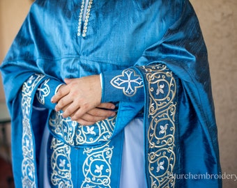 May include: A blue liturgical robe with gold and white embroidery. The robe features a cross design on the sleeve and intricate floral patterns throughout.
