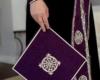 May include: A purple liturgical book with a silver cross design on the cover. The book is held by a person wearing a purple robe with silver trim.