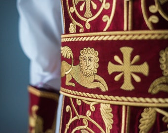 May include: Close-up of a red fabric with gold embroidery. The fabric features a lion, a cross, and floral designs.